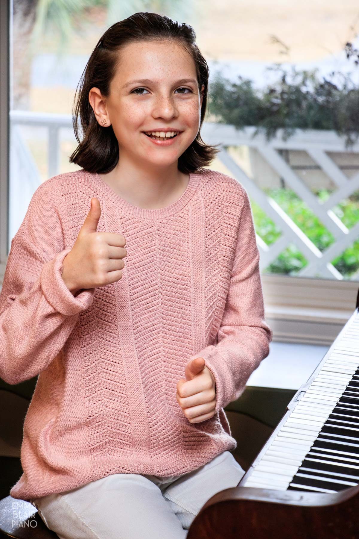 young girl smiling, sitting at the piano, and doing thumbs up